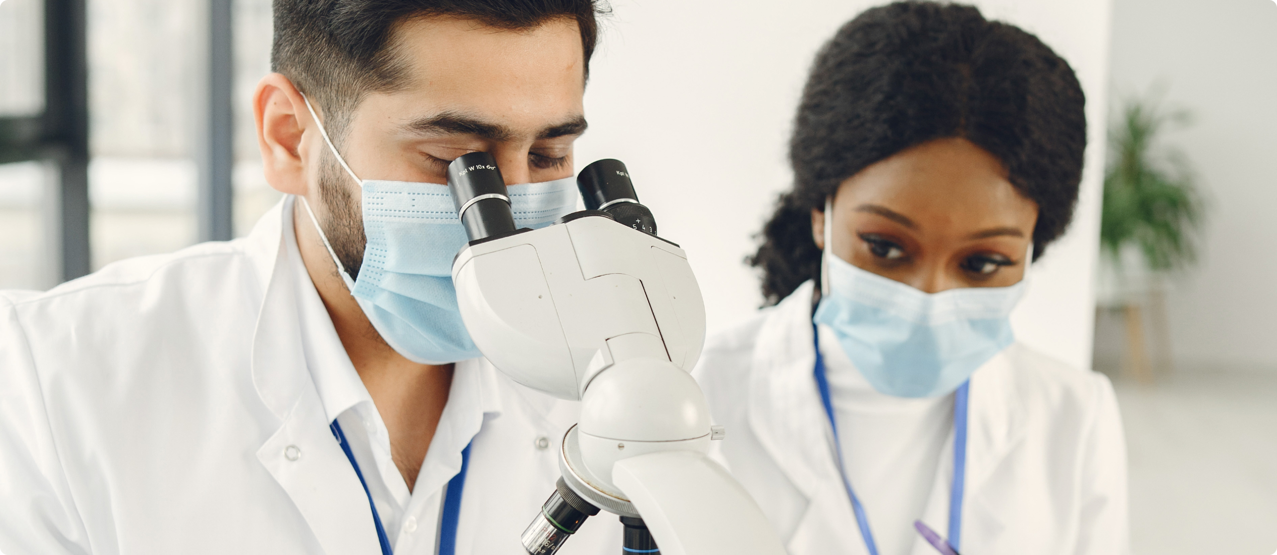 Two masked lab professionals in white coats peer into a microscope in a bright laboratory.