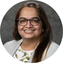 Portrait of a smiling woman with long dark hair and glasses, wearing a white floral blouse, against a gray studio background; warm, approachable tone.