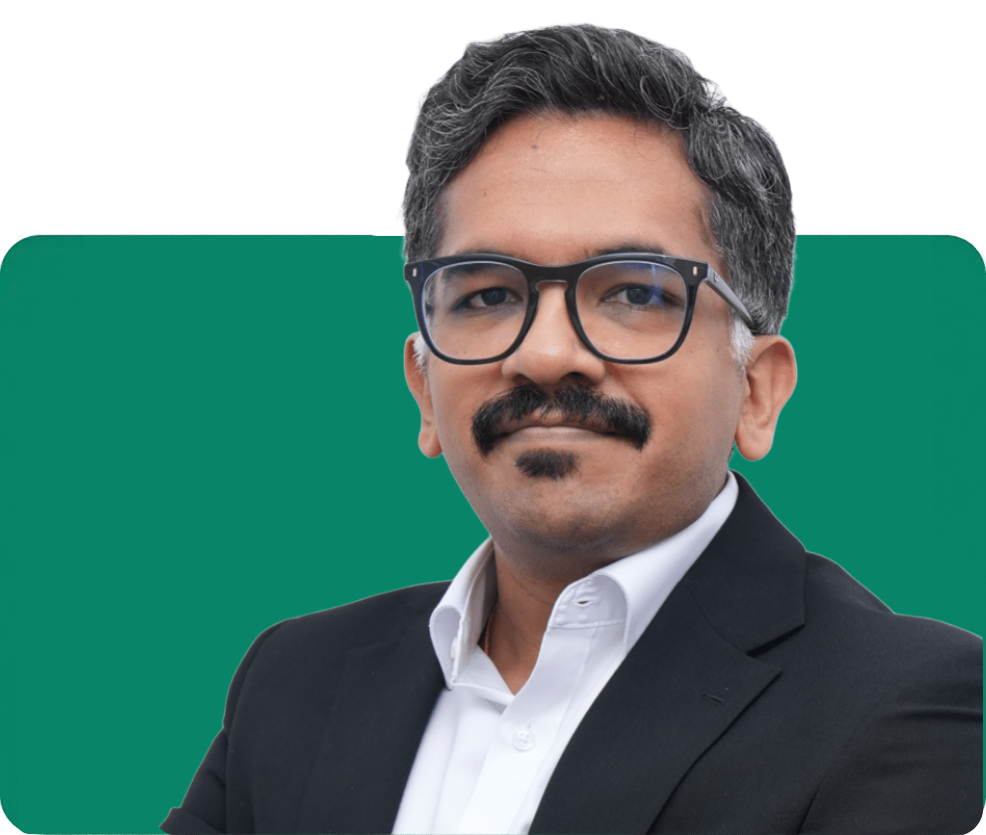 Portrait of a mustached man wearing black glasses, a dark suit, and white shirt against a green panel and white background; he looks calm and confident.