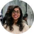 A circular portrait of a smiling woman with curly dark hair and glasses, wearing a light-colored top in a softly blurred modern indoor setting; warm, friendly mood.