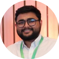 Smiling man with glasses and a beard, wearing a white shirt and green lanyard, photographed against a warm pink curtain background in a circular frame.